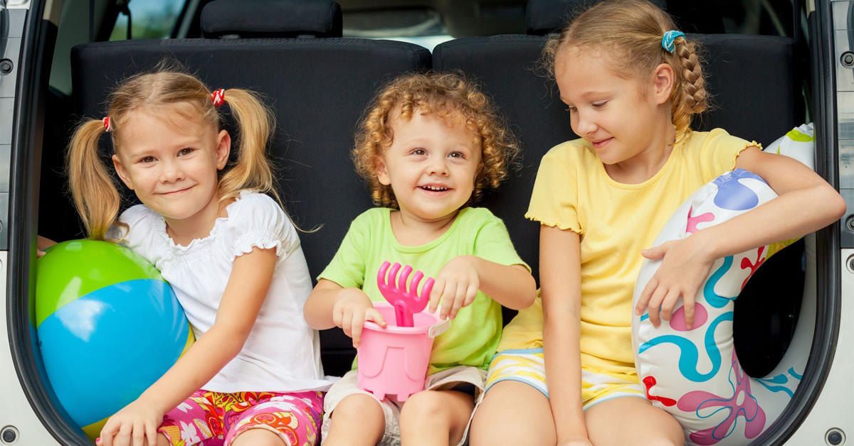 Drie kinderen met strandspullen zitten aan de rand van de achterbak in een auto